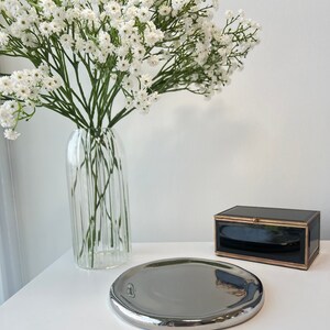 May include: A silver, round, decorative tray with a slightly raised edge. The tray is sitting on a white surface next to a black box with gold trim.