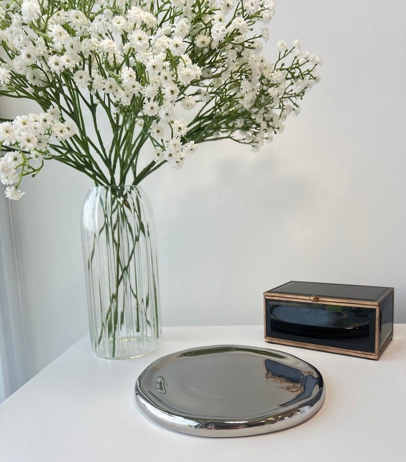 May include: A silver, round, decorative tray with a slightly raised edge. The tray is sitting on a white surface next to a black box with gold trim.