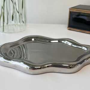 May include: A silver, irregularly shaped tray with a smooth, reflective surface. The tray is made of metal and has a modern, abstract design.