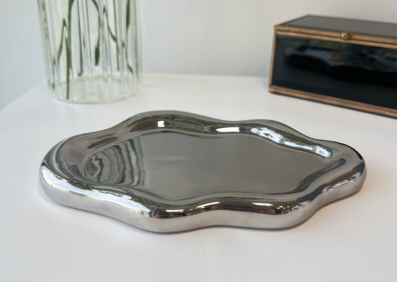 May include: A silver, irregularly shaped tray with a smooth, reflective surface. The tray is made of metal and has a modern, abstract design.