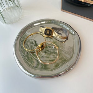 May include: A silver tray with a marbled design in shades of green and white. The tray features a gold watch, a gold bracelet, and a gold ring.