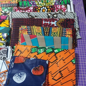 May include: A stack of colorful fabric scraps with various patterns, including geometric, floral, and abstract designs. The fabrics are arranged in a vertical stack, with the top fabric being a dark blue with white and orange designs.