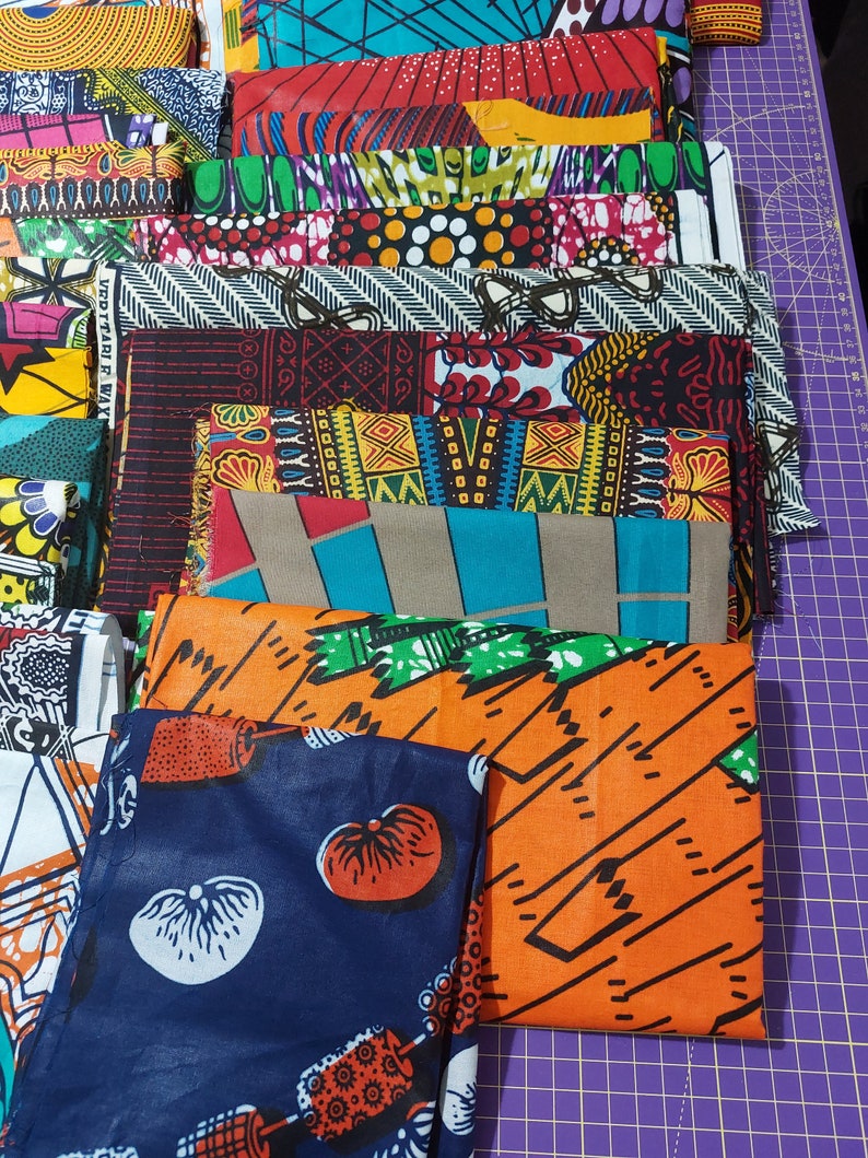 May include: A stack of colorful fabric scraps with various patterns, including geometric, floral, and abstract designs. The fabrics are arranged in a vertical stack, with the top fabric being a dark blue with white and orange designs.