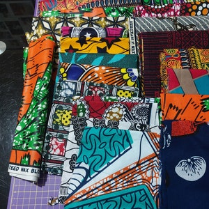 May include: A collection of colorful fabric scraps with various patterns, including geometric, floral, and abstract designs. The fabrics are arranged in a stack and labeled "STEED WAX BLOCK".