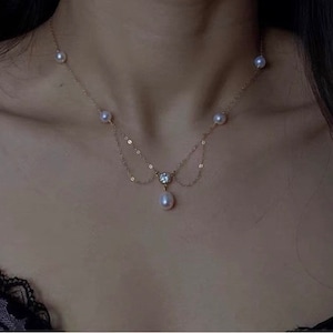 Puede incluir: Un delicado collar de oro con una serie de pequeñas perlas y un colgante central. El colgante incluye una pequeña gema transparente y una perla más grande en forma de lágrima. El collar está diseñado para ser usado alrededor del cuello.
