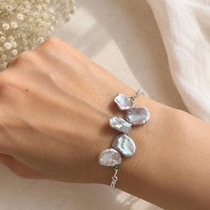 May include: A delicate bracelet featuring irregularly shaped, iridescent white pearls. The pearls are connected by a fine silver chain and accented with small green beads. The bracelet is shown on a wrist, showcasing its elegant design.