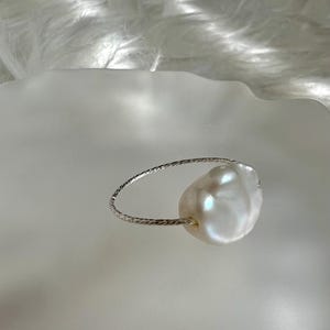 May include: A silver ring with a large, white baroque pearl. The ring is made of a thin, twisted wire.