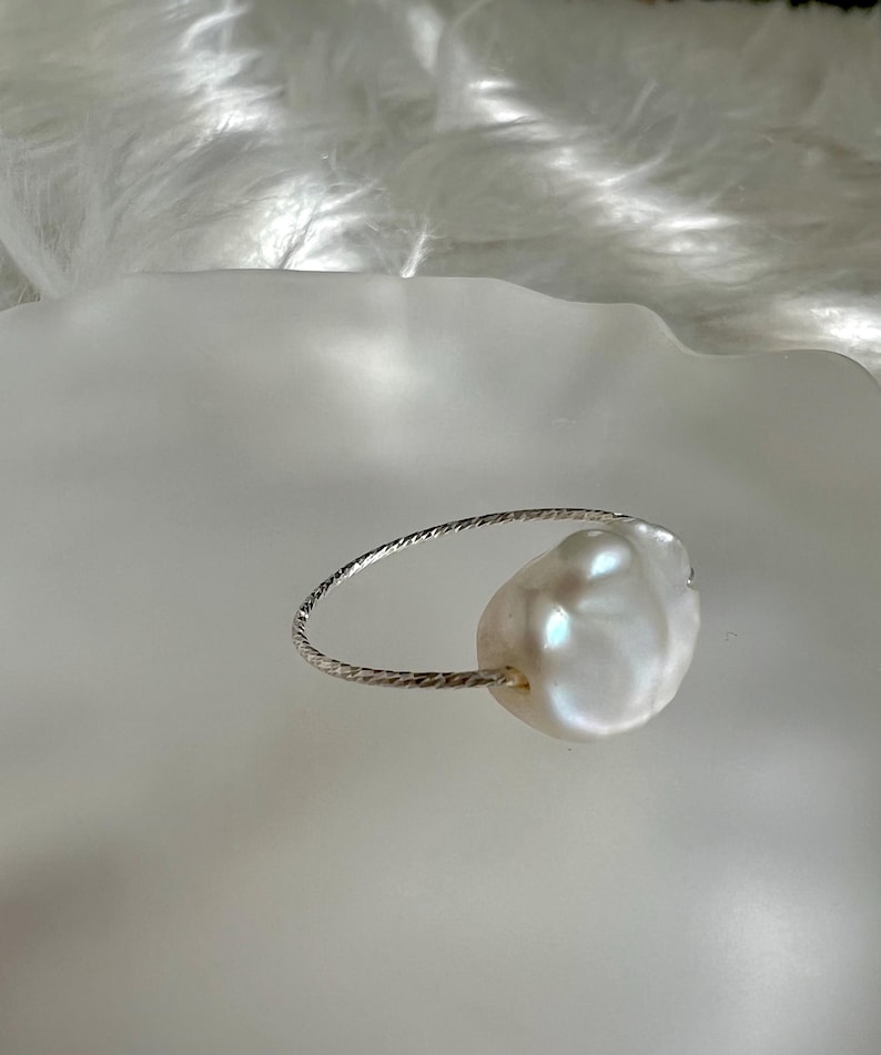 May include: A silver ring with a large, white baroque pearl. The ring is made of a thin, twisted wire.