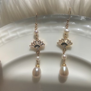 Pode incluir: Um par de brincos de ouro com um design em forma de leque com strass transparentes e uma pérola pendurada.