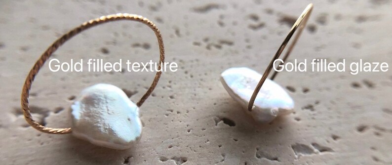 May include: Two gold filled rings with a white pearl. One ring has a textured band and the other has a smooth band. The text "Gold filled texture" is above the ring with the textured band. The text "Gold filled glaze" is above the ring with the smooth band.