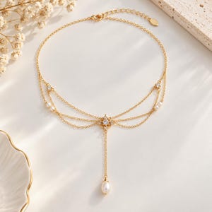 Puede incluir: Un collar dorado de varias cadenas finas, peque&ntilde;as perlas blancas y piedras transparentes brillantes. El dise&ntilde;o incluye un colgante central con forma de flor del que cuelga una perla en forma de gota, con cierre de mosquet&oacute;n.