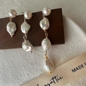 Puede incluir: Un par de pendientes de gota de perla con una cadena de oro y múltiples acentos de perla. Los pendientes se muestran sobre una superficie de madera con un fondo blanco.