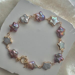 Puede incluir: Un brazalete de cadena dorada con dijes de perlas en forma de estrella en tonos blanco, rosa y morado.