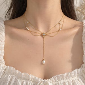 Puede incluir: Un collar dorado de varias hebras con cadenas delicadas dispuestas en un dise&ntilde;o superpuesto. El centro presenta un peque&ntilde;o colgante en forma de estrella con una piedra clara, acentuado por peque&ntilde;as perlas blancas y piedras claras adicionales.