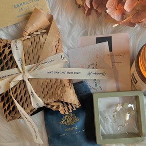 May include: A pair of white and green gemstone earrings in a small gray box. The earrings are on a white fuzzy surface with a candle, a brown paper package, and a blue pouch with a lotus flower logo.