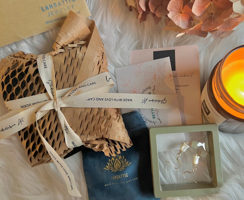 May include: A pair of white and green gemstone earrings in a small gray box. The earrings are on a white fuzzy surface with a candle, a brown paper package, and a blue pouch with a lotus flower logo.