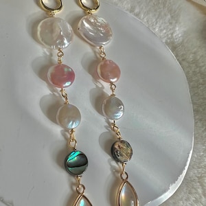 Pode incluir: Um par de brincos longos de aro dourado com vários pingentes de pérola e concha. Os brincos apresentam uma pérola branca grande, uma pérola rosa menor, uma pérola branca, uma concha de abalone verde e marrom e uma pérola iridescente em forma de lágrima.