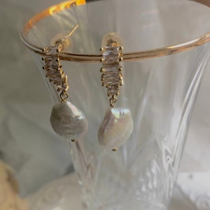 Pode incluir: Brincos de ouro com uma p&eacute;rola pendurada e uma fileira de pedras de zirc&ocirc;nia c&uacute;bica transparentes.
