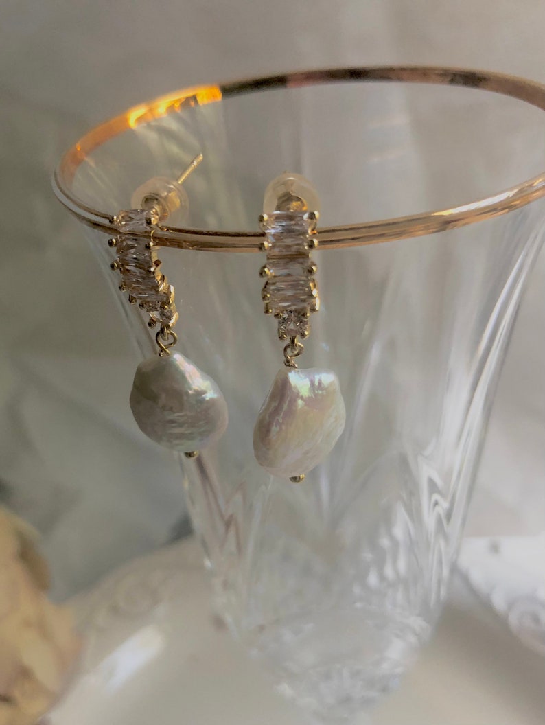 Pode incluir: Brincos de ouro com uma p&eacute;rola pendurada e uma fileira de pedras de zirc&ocirc;nia c&uacute;bica transparentes.