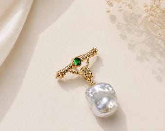 Puede incluir: Un broche de tono dorado con una barra horizontal de textura retorcida, una piedra preciosa verde redonda en el centro y una perla barroca blanca irregular colgante. La barra de metal está decorada con pequeñas piedras claras.
