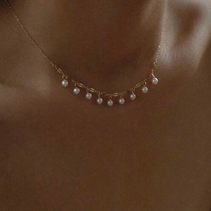 Puede incluir: Un delicado collar de cadena dorada con pequeñas perlas blancas que cuelgan. El collar lo lleva una persona de tez morena clara.
