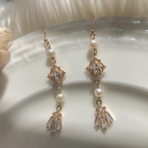 Pode incluir: Brincos de argola dourados com um cristal quadrado e detalhes de pérolas. Os brincos têm um design delicado e feminino.