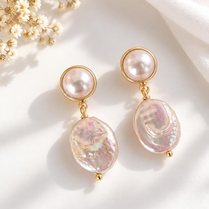 Pode incluir: Um par de brincos pendentes com uma pérola redonda, lisa e rosa-pálido, engastada numa moldura de metal dourado, ligada por um pequeno elo dourado a uma pérola barroca plana, irregular e iridescente, com uma pequena conta dourada na base.