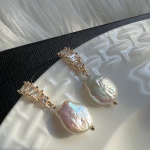 Pode incluir: Um par de brincos de ouro com uma gota de p&eacute;rola e uma fileira de cristais transparentes em forma de baguette.