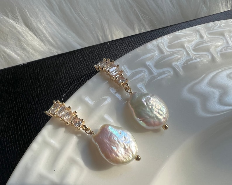 Pode incluir: Um par de brincos de ouro com uma gota de p&eacute;rola e uma fileira de cristais transparentes em forma de baguette.