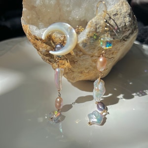 Pode incluir: Um par de brincos longos e pendentes com um amuleto de lua crescente, contas de vidro iridescentes e pérolas barrocas. Os brincos são feitos de metal dourado.