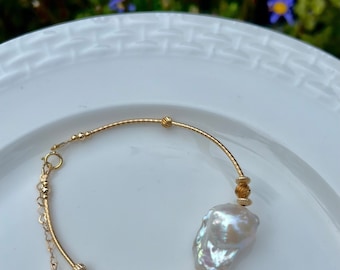 Peut inclure: Un bracelet en or avec une grosse perle irrégulière et une petite perle pendante sur une chaîne. Le bracelet est posé sur une assiette blanche.