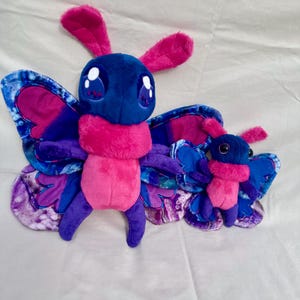 Puede incluir: Dos juguetes de peluche de mariposa. La mariposa más grande tiene cabeza azul, cuerpo rosa y patas moradas. Sus alas son azules y rosas con un diseño veteado. La mariposa más pequeña tiene características similares.