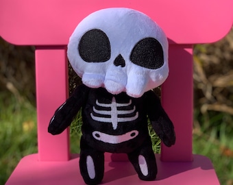 May include: A plush skeleton toy with a white skull and black body. The toy is sitting on a pink chair.