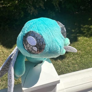 May include: A plush toy featuring a bright turquoise body with large, round eyes that have a silver, honeycomb-patterned iris and white pupils. The toy has two prominent, flat, white wings extending from its sides, decorated with a black grid pattern.