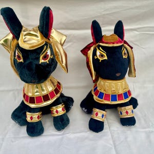 Op de afbeelding: Twee pluche Anubis-figuren. De zwarte pluche honden hebben gouden, rode en blauwe decoratieve kragen, armbanden en hoofddeksels. De Anubis-figuren hebben grote oren en zitten rechtop. De achtergrond is een effen wit oppervlak.