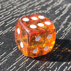 May include: A transparent orange dice with white dots, resting on a dark, textured surface. The dice is lit by sunlight, casting a shadow. The dice shows the number six.