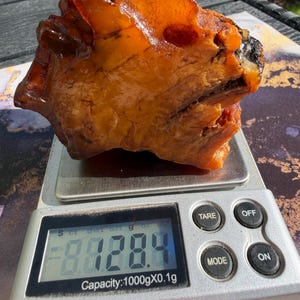 Puede incluir: Una pieza grande e irregular de ámbar translúcido con tonos naranja intenso y dorados, que presenta fracturas internas visibles y una superficie lisa y pulida. El ámbar descansa sobre una báscula digital que muestra 128,4 gramos.