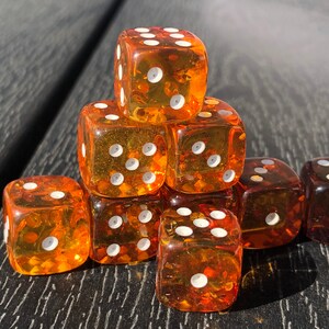 May include: A collection of transparent amber-colored dice with white pips. The dice are stacked in a pyramid shape, with some dice scattered around. The dice are on a dark wooden surface, creating a contrast between the dice and the background.