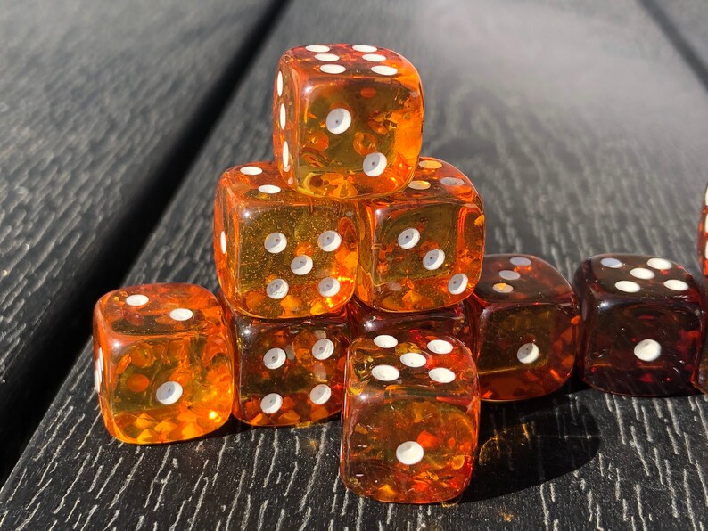 May include: A collection of transparent amber-colored dice with white pips. The dice are stacked in a pyramid shape, with some dice scattered around. The dice are on a dark wooden surface, creating a contrast between the dice and the background.