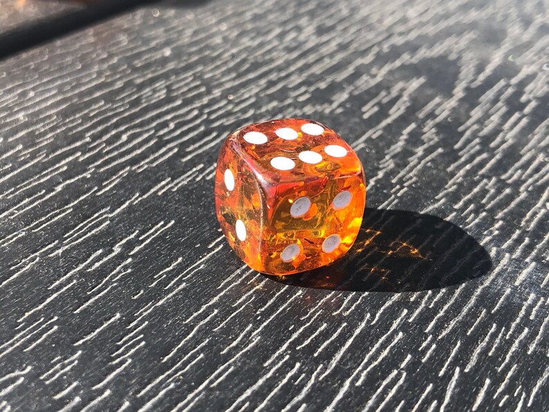 May include: A transparent orange dice with white dots, resting on a dark, textured surface. The dice is lit by sunlight, casting a shadow. The dice shows the number six.