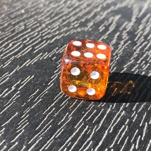 May include: A transparent orange die with white dots, showing the number six. The die is resting on a dark, textured surface. The image is lit by sunlight, casting a shadow.