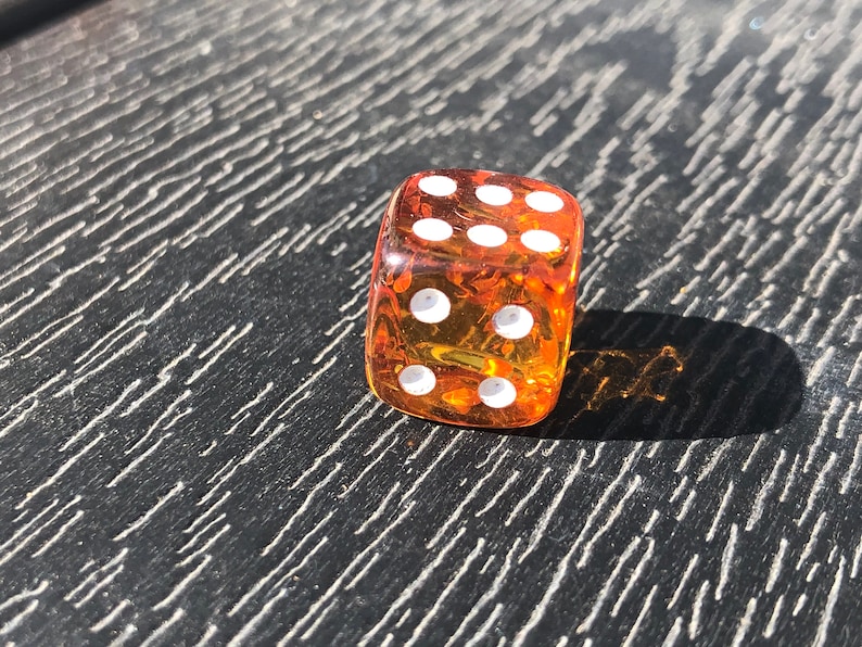 May include: A transparent orange die with white dots, showing the number six. The die is resting on a dark, textured surface. The image is lit by sunlight, casting a shadow.