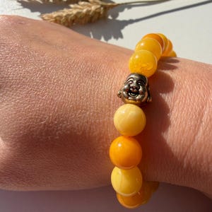 May include: A beaded bracelet featuring amber-colored beads and a silver-colored Buddha charm. The bracelet is composed of various shades of yellow and orange beads. The bracelet is worn on a wrist.