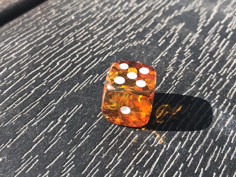 May include: A transparent amber-colored die with white dots, showing the number six. The die is resting on a dark, textured surface, casting a shadow. The image is taken in bright sunlight.