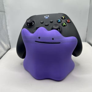 Puede incluir: Un soporte para mando de color morado con forma de mancha amorfa, que presenta dos peque&ntilde;os puntos negros como ojos y una fina l&iacute;nea negra horizontal como boca, situado delante de un mando de juego inal&aacute;mbrico negro con botones de colores.