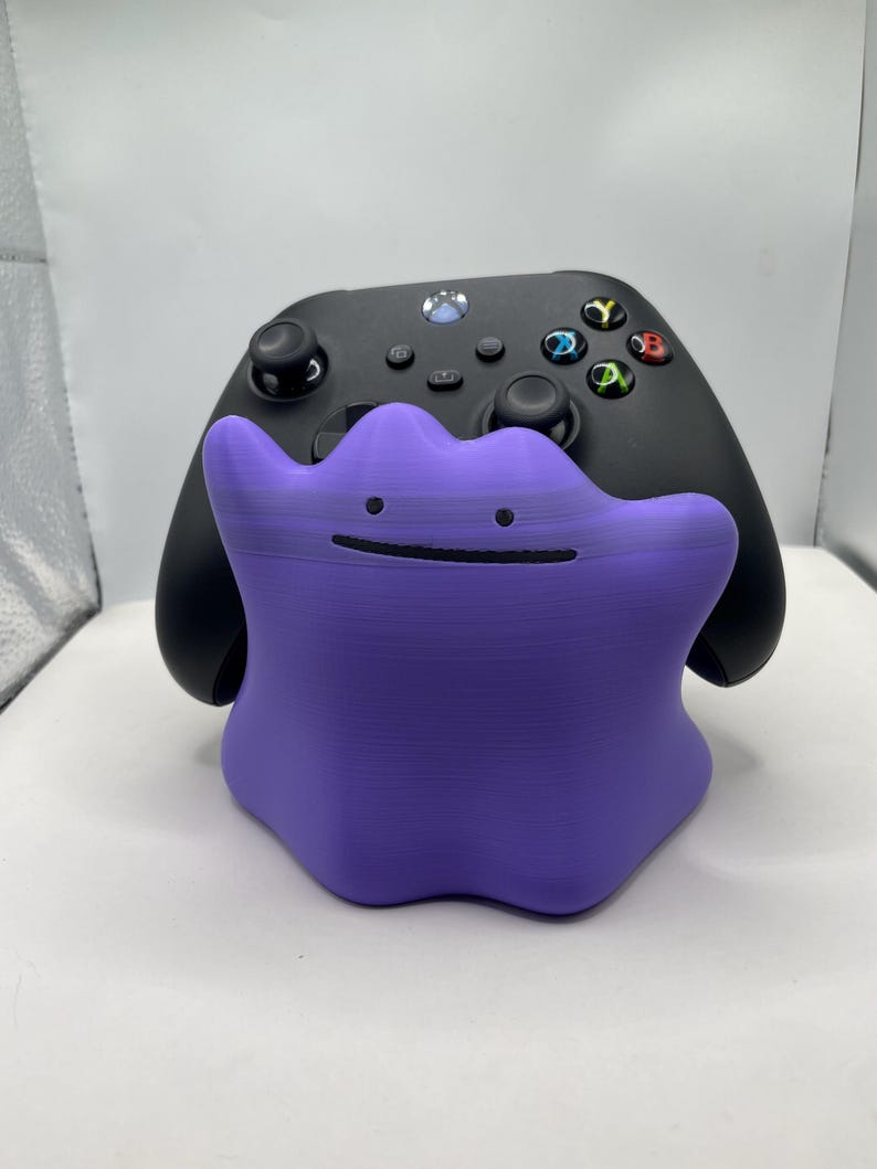 Puede incluir: Un soporte para mando de color morado con forma de mancha amorfa, que presenta dos peque&ntilde;os puntos negros como ojos y una fina l&iacute;nea negra horizontal como boca, situado delante de un mando de juego inal&aacute;mbrico negro con botones de colores.