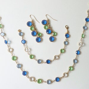 K&ouml;nnte beinhalten: Ein Schmuckset mit einer Halskette, einem Armband und Ohrringen. Der Schmuck besteht aus goldenen Ketten mit blauen, gr&uuml;nen und klaren runden Glasperlen.