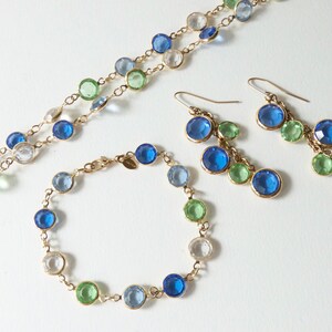K&ouml;nnte beinhalten: Ein Schmuckset mit einer Halskette, einem Armband und Ohrringen. Die St&uuml;cke bestehen aus einer goldenen Kette und haben blaue, gr&uuml;ne und klare Glasperlen.