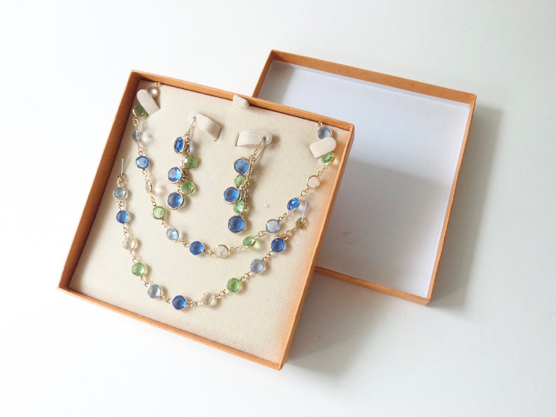 K&ouml;nnte beinhalten: Ein Set mit blauen und gr&uuml;nen Edelsteinohrringen und einer Halskette in einer beigefarbenen Schmuckschatulle.