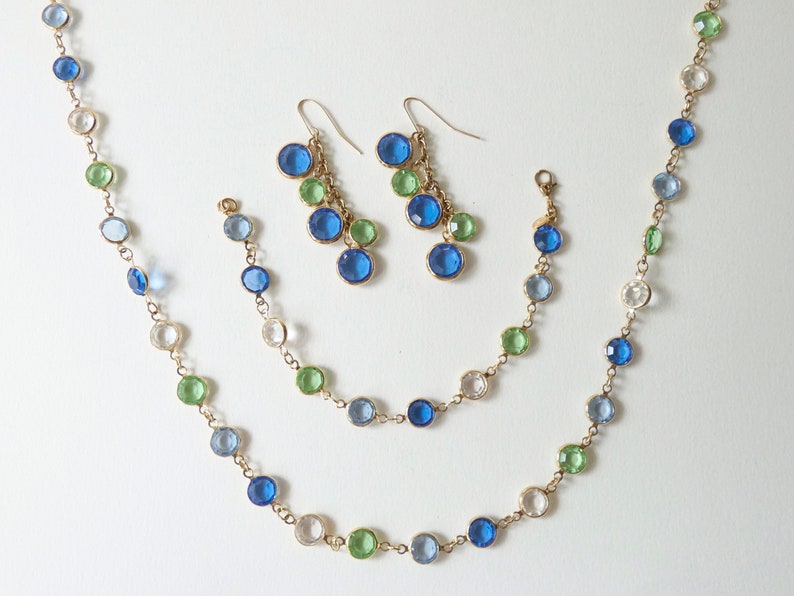 K&ouml;nnte beinhalten: Ein Schmuckset mit einer Halskette, einem Armband und Ohrringen. Der Schmuck besteht aus goldenen Ketten mit blauen, gr&uuml;nen und klaren runden Glasperlen.